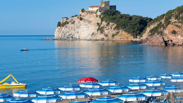 A coastal scene with a historic building on a cliff, calm sea, and rows of blue and white beach umbrellas. - Cosy apartment in villa for 4 guests with WIFI, A/C, pool and terrace (Castiglione Della Pescaia)