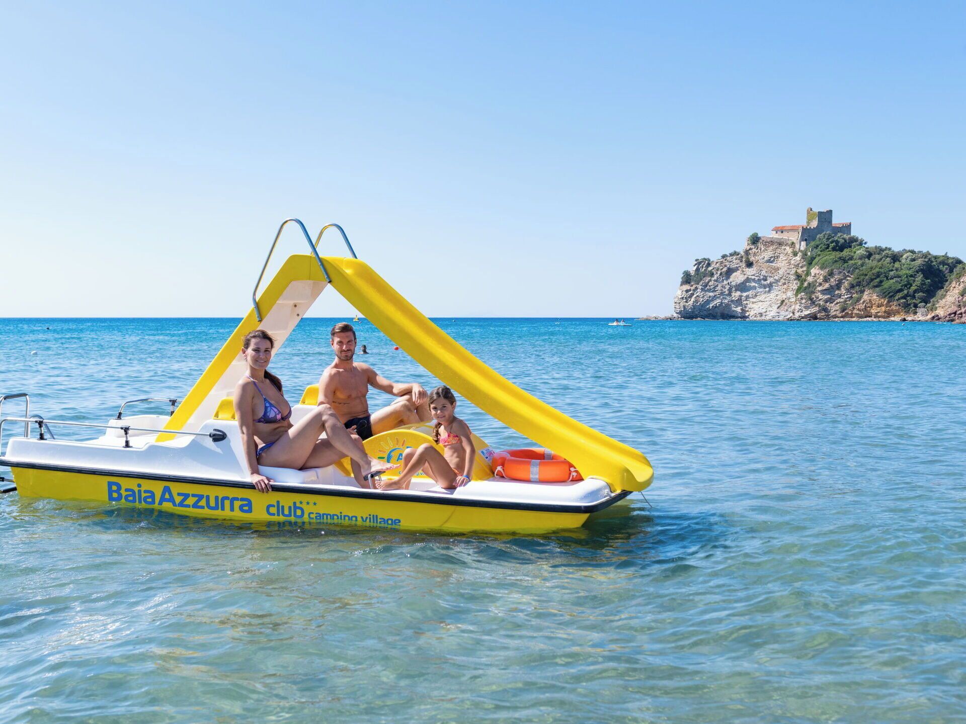 Un barco amarillo y blanco con la leyenda "Baia Azzurra club" y "camping village", flotando sobre aguas tranquilas, con un acantilado rocoso y un castillo al fondo.