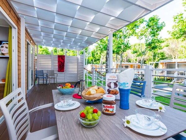 Un espace de restauration en plein air avec une table dressée pour deux personnes, agrémentée d’un saladier de fruits, d’un pot de Nutella, d’un bocal de biscuits et d’une cafetière.