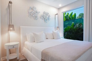 Una camera da letto moderna con un letto grande, biancheria da letto bianca, un comodino, una lampada e vista sul verde attraverso una finestra.