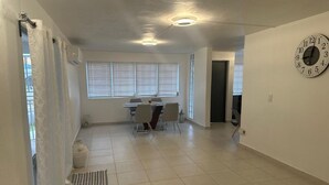 A spacious room with a dining area, a clock on the wall, and a window with blinds.