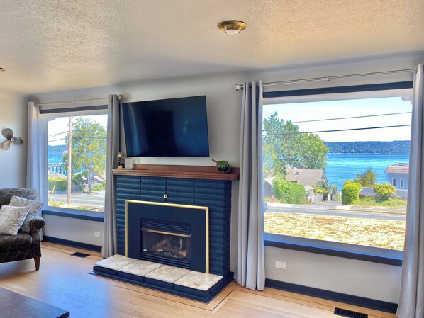 A living room with a fireplace, a flat-screen TV mounted above it, and large windows offering a view of a residential area and a body of water.