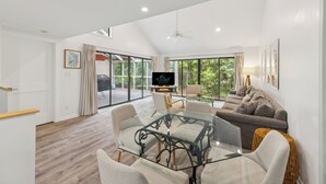 Un salon spacieux avec une table basse en verre, des sièges confortables, un téléviseur à écran plat et de grandes fenêtres donnant sur un paysage verdoyant.