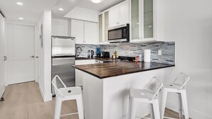 Een moderne keuken met witte kasten, een roestvrijstalen koelkast, een magnetron en een betegelde spatwand. Er staan twee witte barkrukken bij het eiland.