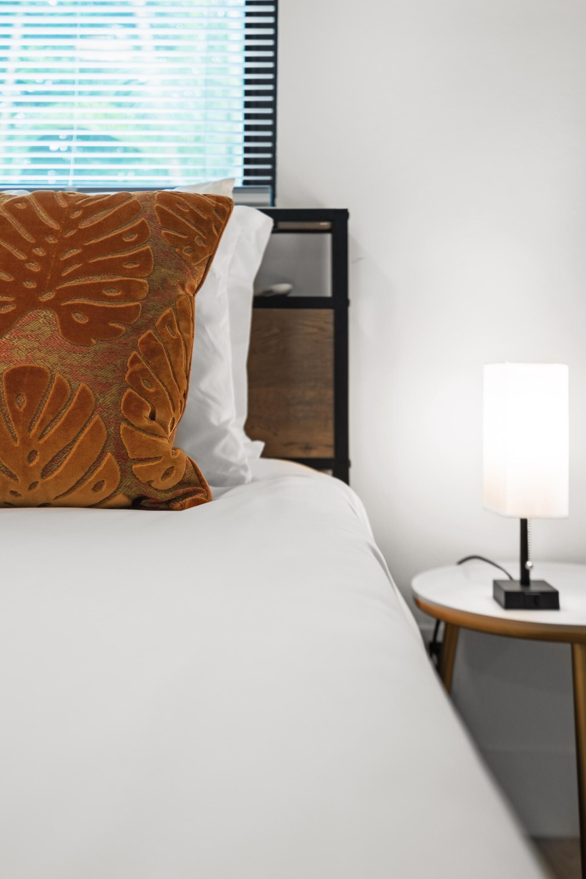 Un lit bien fait, avec un oreiller à motifs blancs et bruns, une table de chevet avec une lampe et une fenêtre avec des stores.