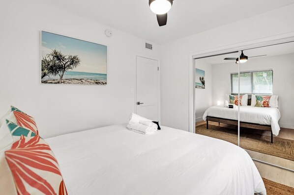 A bedroom with a large bed, a door, a mirror, and a painting of a beach scene.