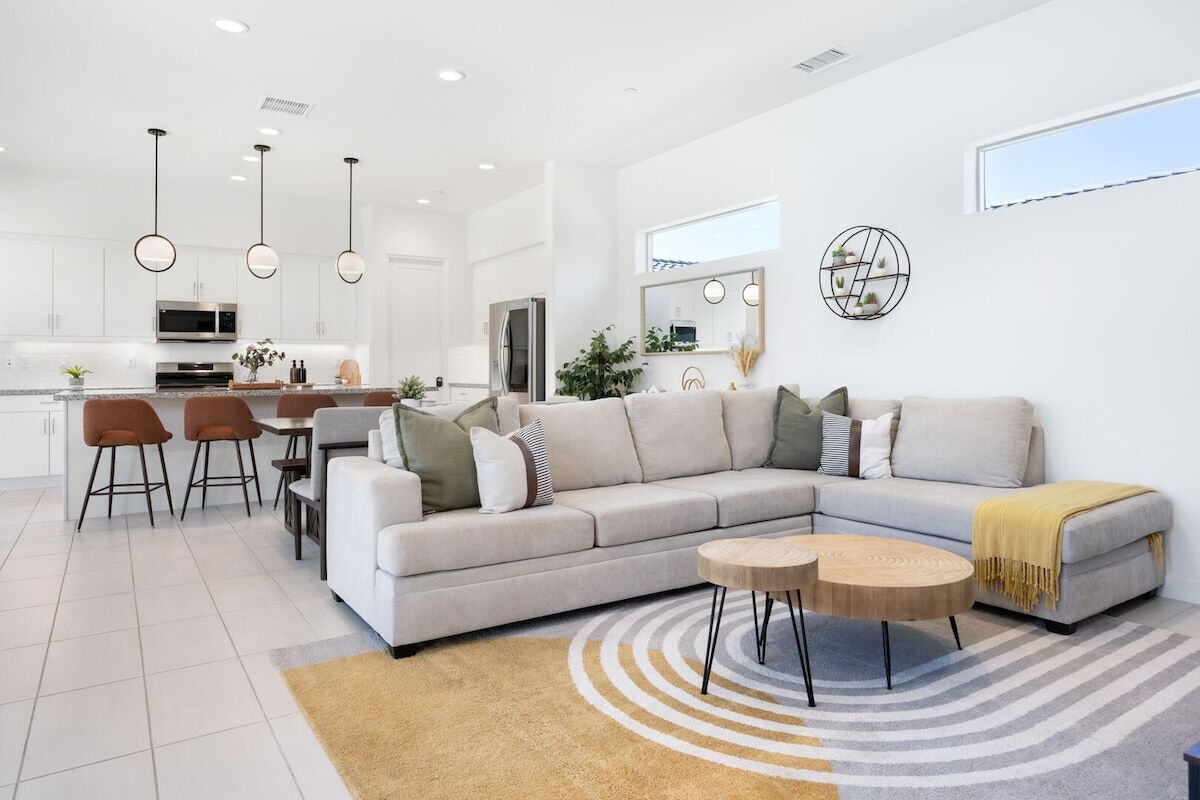 Un salon moderne avec un canapé d’angle, un coin repas avec des chaises, une cuisine en arrière-plan et une grande fenêtre qui laisse entrer la lumière naturelle.