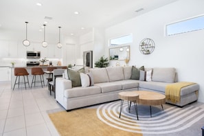 A modern living room with a sectional sofa, a dining area with chairs, a kitchen in the background, and a large window providing natural light. - Play Paradise: Pool+Spa, BBQ, GameRoom, PGA West! (La Quinta)