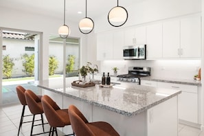 A modern kitchen with white cabinets, a granite countertop, and a central island with bar stools. - Play Paradise: Pool+Spa, BBQ, GameRoom, PGA West! (La Quinta)