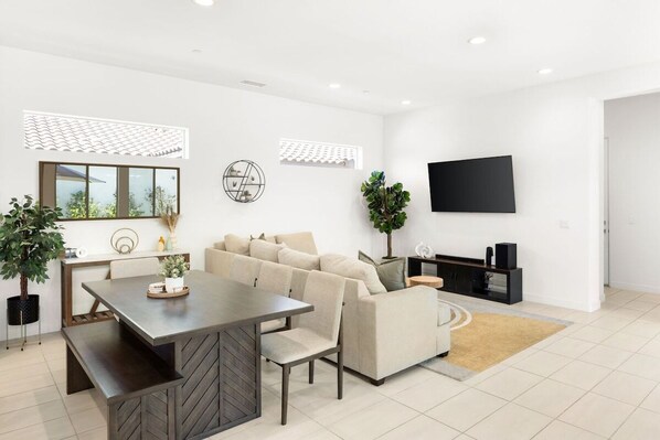 A modern living room with a sofa, a dining table, and a TV mounted on the wall. - Play Paradise: Pool+Spa, BBQ, GameRoom, PGA West! (La Quinta)