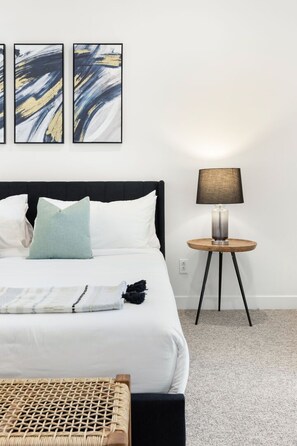 En pænt redt seng med hvidt sengetøj, en enkelt blå pude og et sort plaide. Der er et rundt træsidebord med en lampe ved siden af sengen.