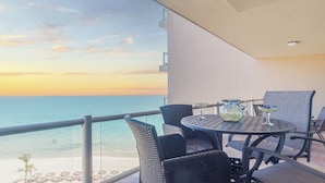 Een balkon met uitzicht op zee tijdens de zonsondergang, voorzien van rieten stoelen en een ronde tafel met glazen.
