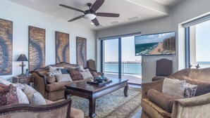 Un ampio soggiorno con una grande finestra che offre vista sull'oceano e sulla città. Presente una ventola a soffitto, una televisione e un tavolino da caffè con soprammobili.