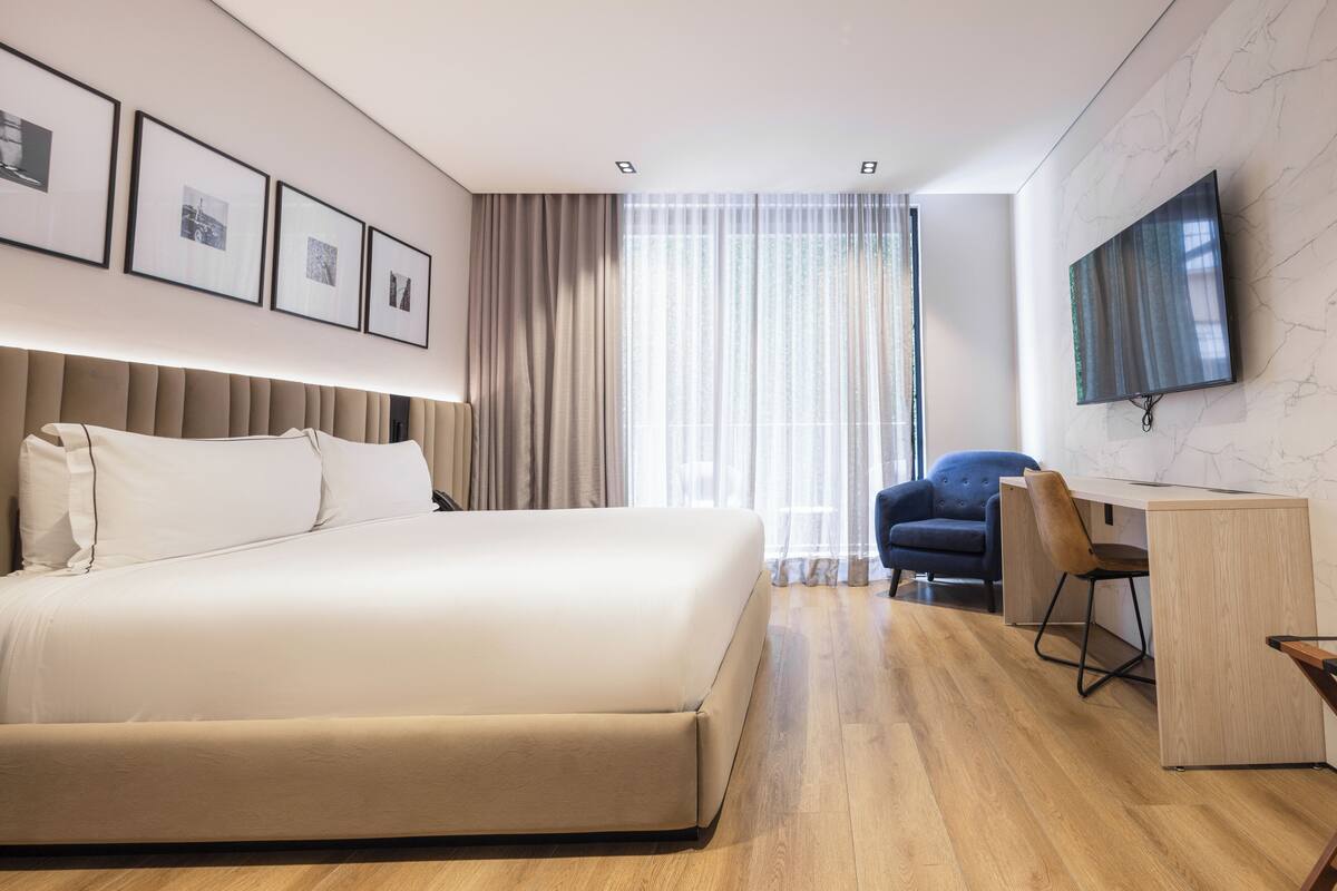 Una habitación de hotel moderna con una cama grande, un escritorio con una silla, un televisor de pantalla plana montado en la pared y un amplio ventanal con cortinas translúcidas.