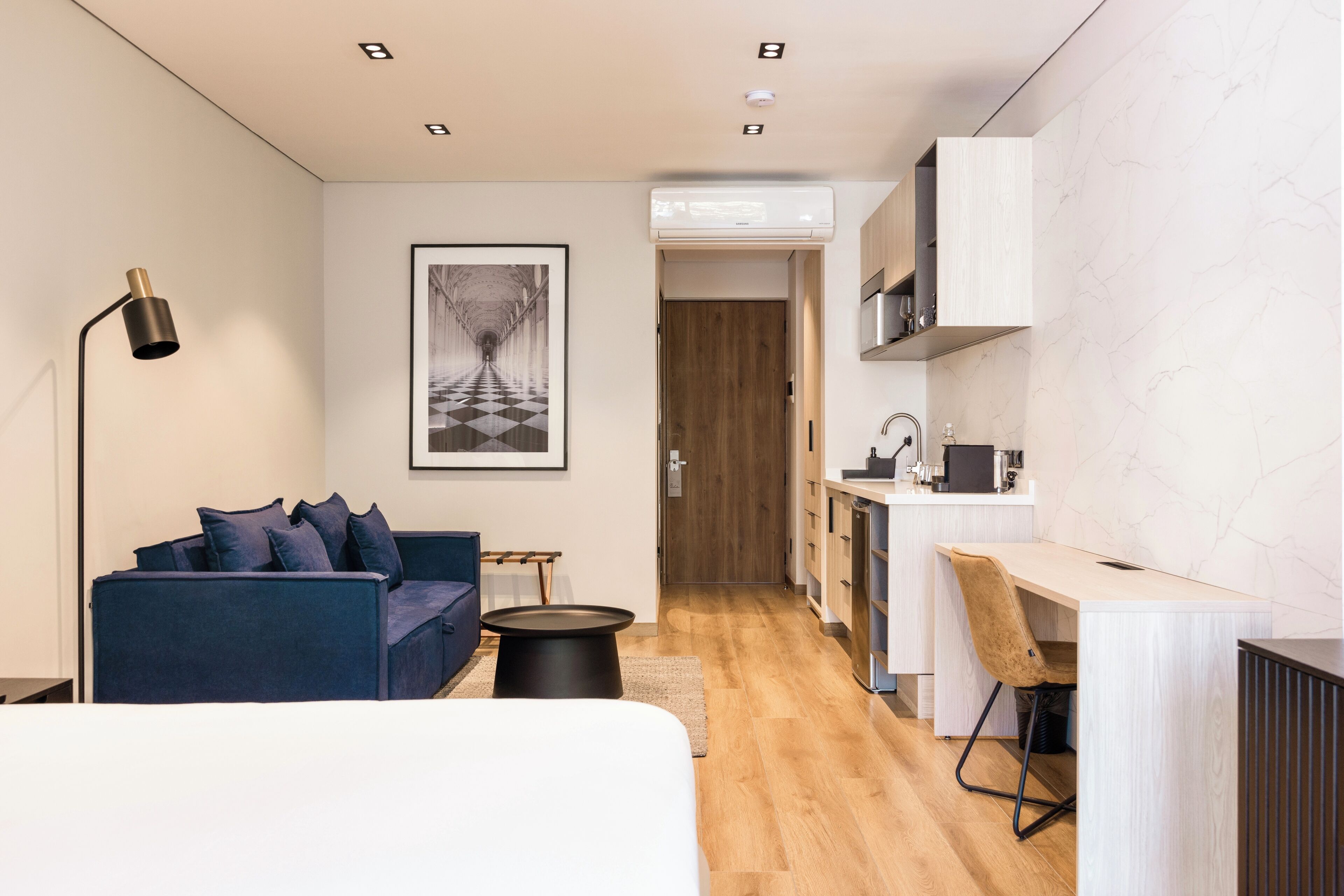 A modern hotel room with a blue sofa, a small round table, a desk with a chair, and a kitchenette.