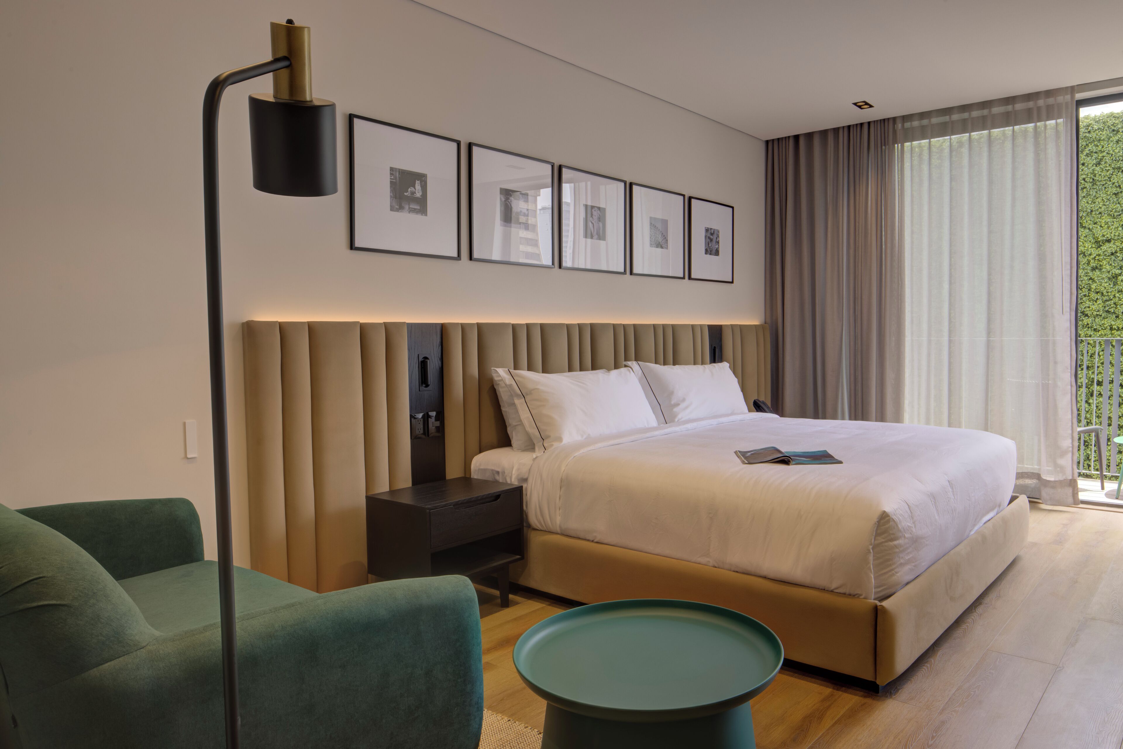 A modern hotel room with a large bed, a bedside table, a green armchair, a floor lamp, and a view of greenery through the window.