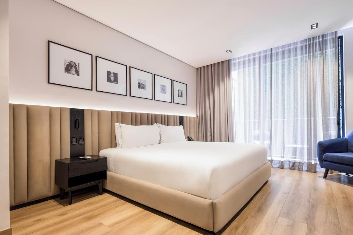 Un dormitorio moderno con una cama grande, una mesita de noche, una silla y una ventana con cortinas traslúcidas.