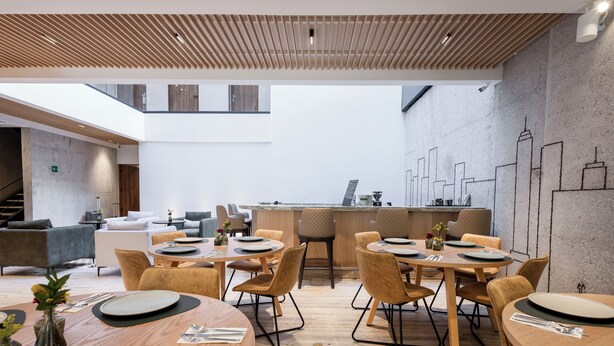 Een moderne eetruimte met houten tafels en stoelen, een keuken op de achtergrond en een stadsgezicht muurschildering op de wand.