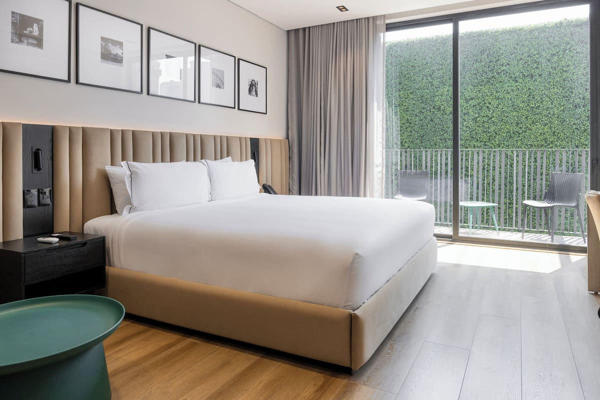 Habitación de hotel moderna con una cama grande, una mesita de noche, una pequeña mesa verde y un balcón con vistas a la vegetación.
