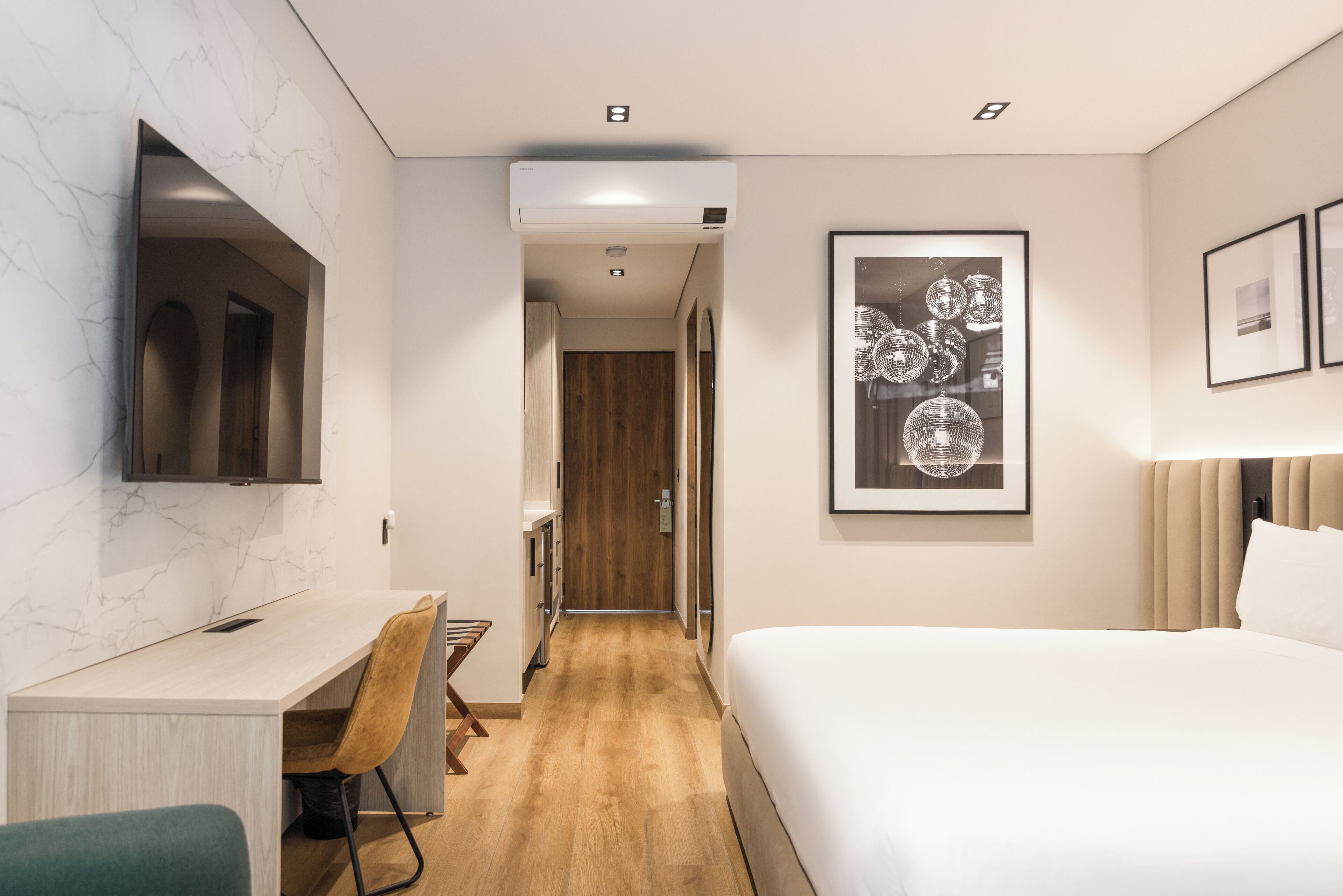 A modern hotel room with a wooden floor, a desk with a chair, a bed, a TV mounted on the wall, and framed artwork.