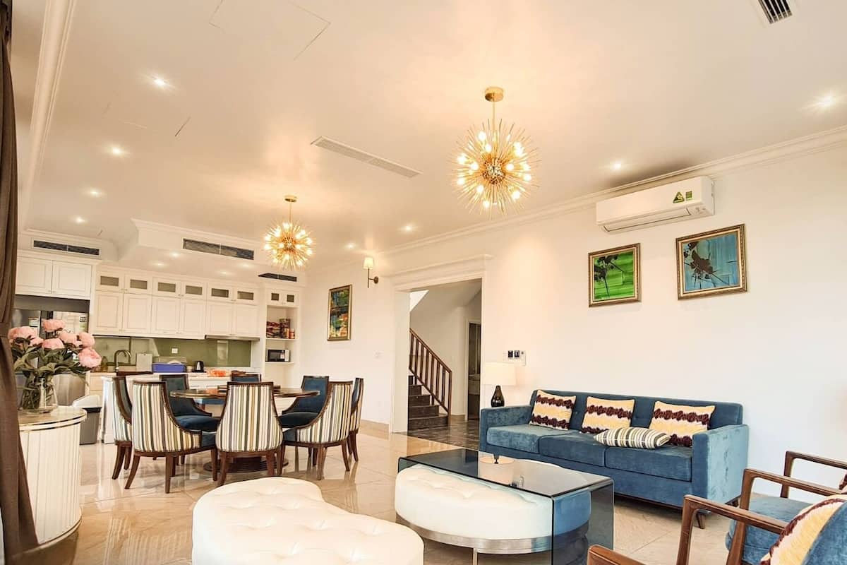 A modern living room with a blue sofa, a glass coffee table, striped chairs, and a chandelier. There is a kitchen in the background with white cabinets and a dining area with a table and chairs.