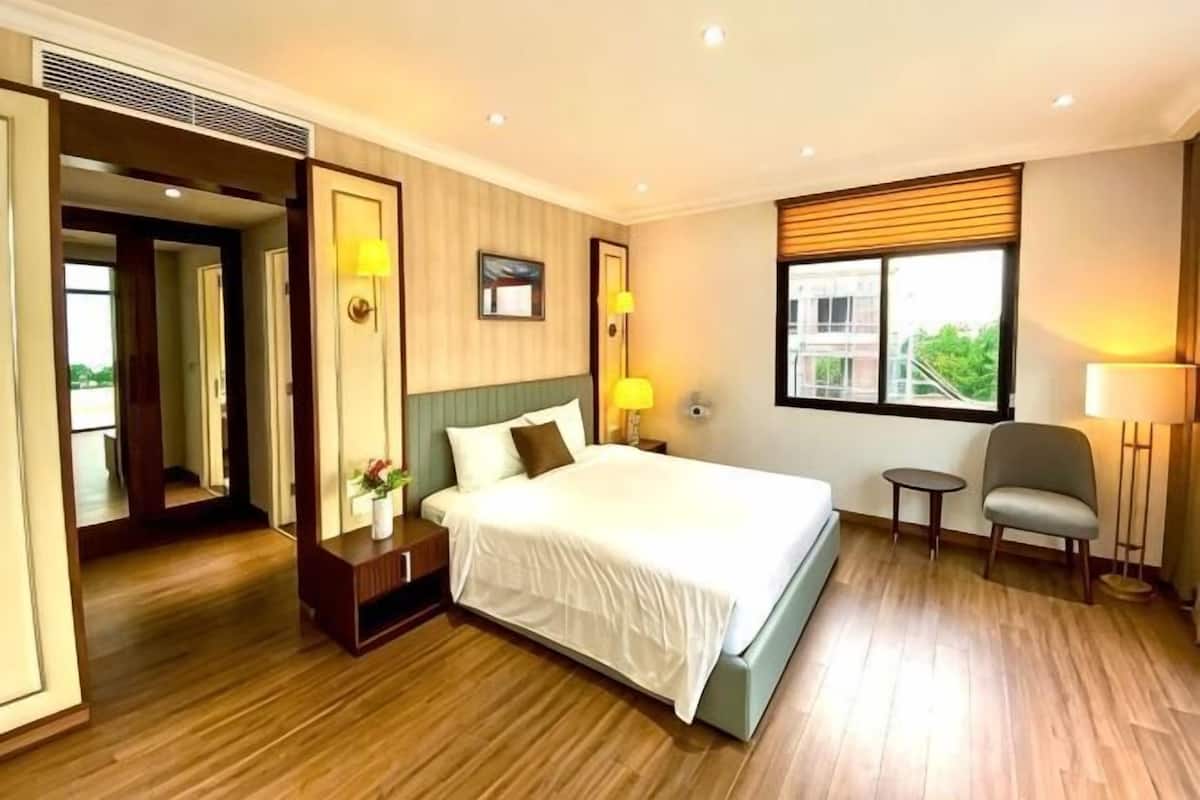 A hotel room with a large bed, bedside tables, a chair, a window with blinds, and a door leading to another room.