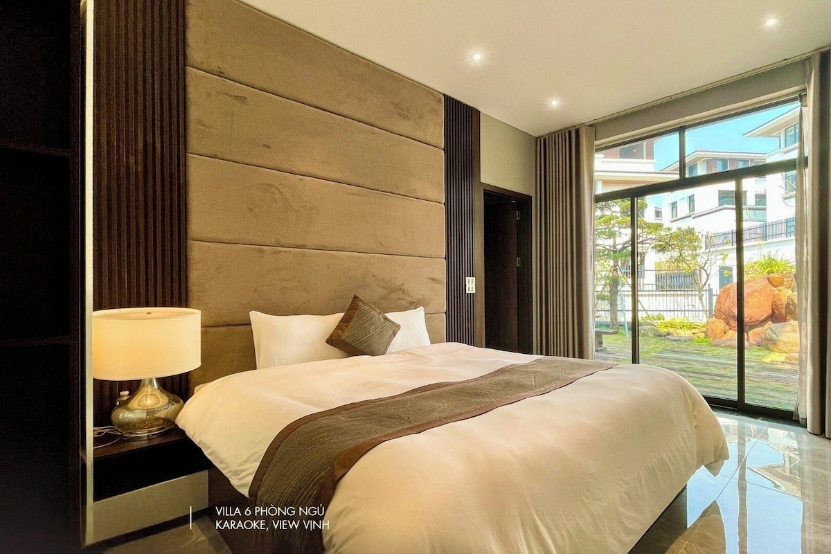 A modern hotel room with a large bed, a nightstand, a lamp, and a view of the outside.