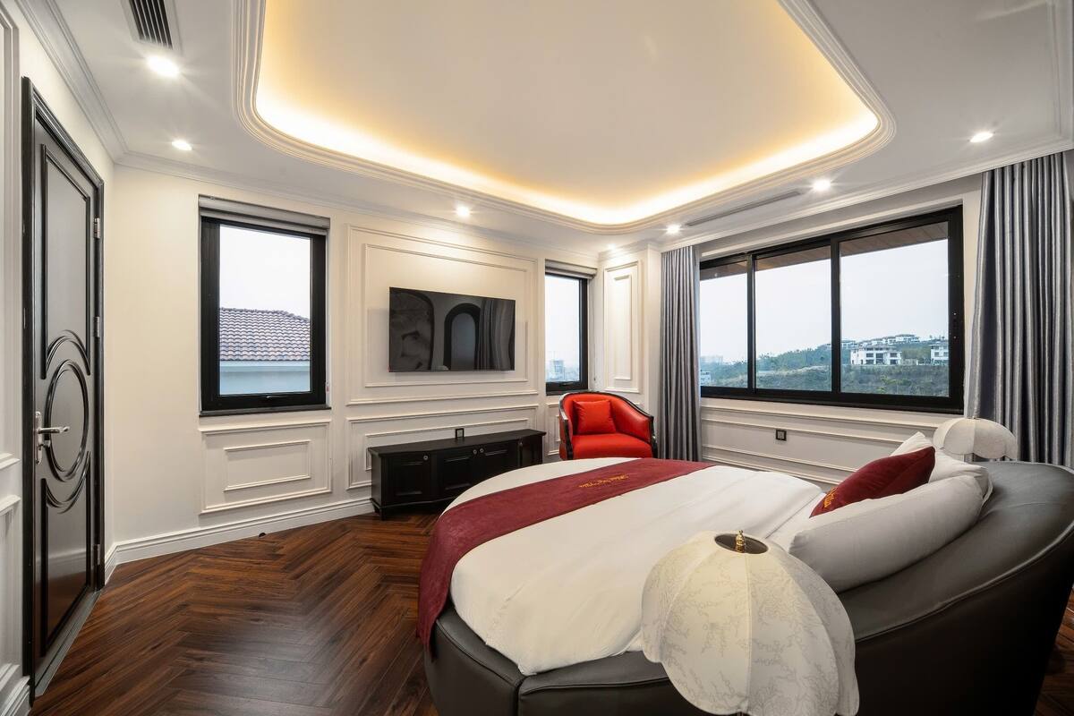 A modern hotel room with a large bed, a red chair, a fireplace, and large windows with a city view.