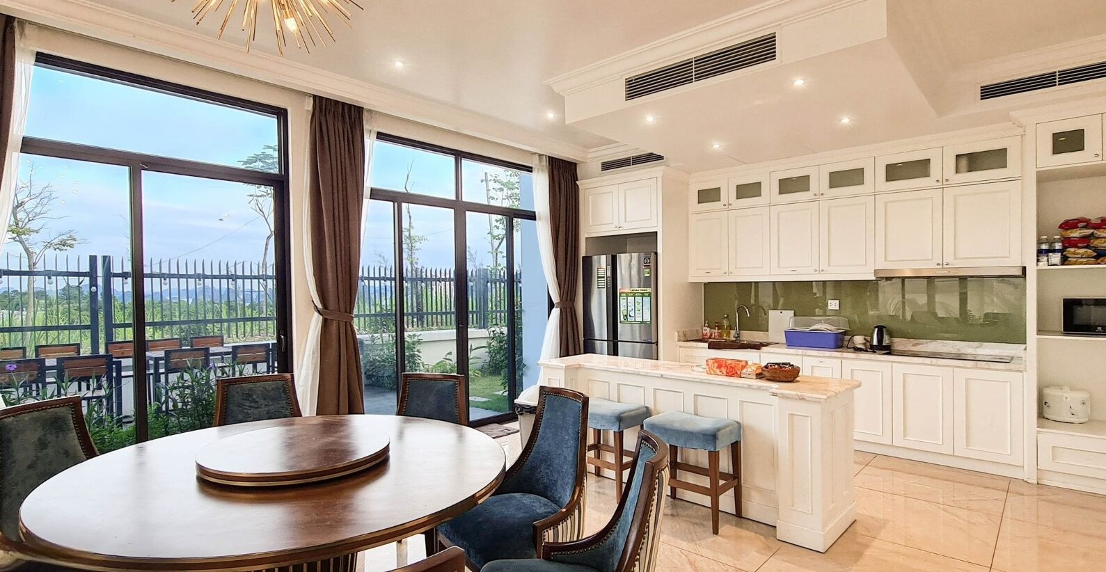 A dining area with a round table and chairs, a kitchen with white cabinets and a large window, and a view of greenery outside.