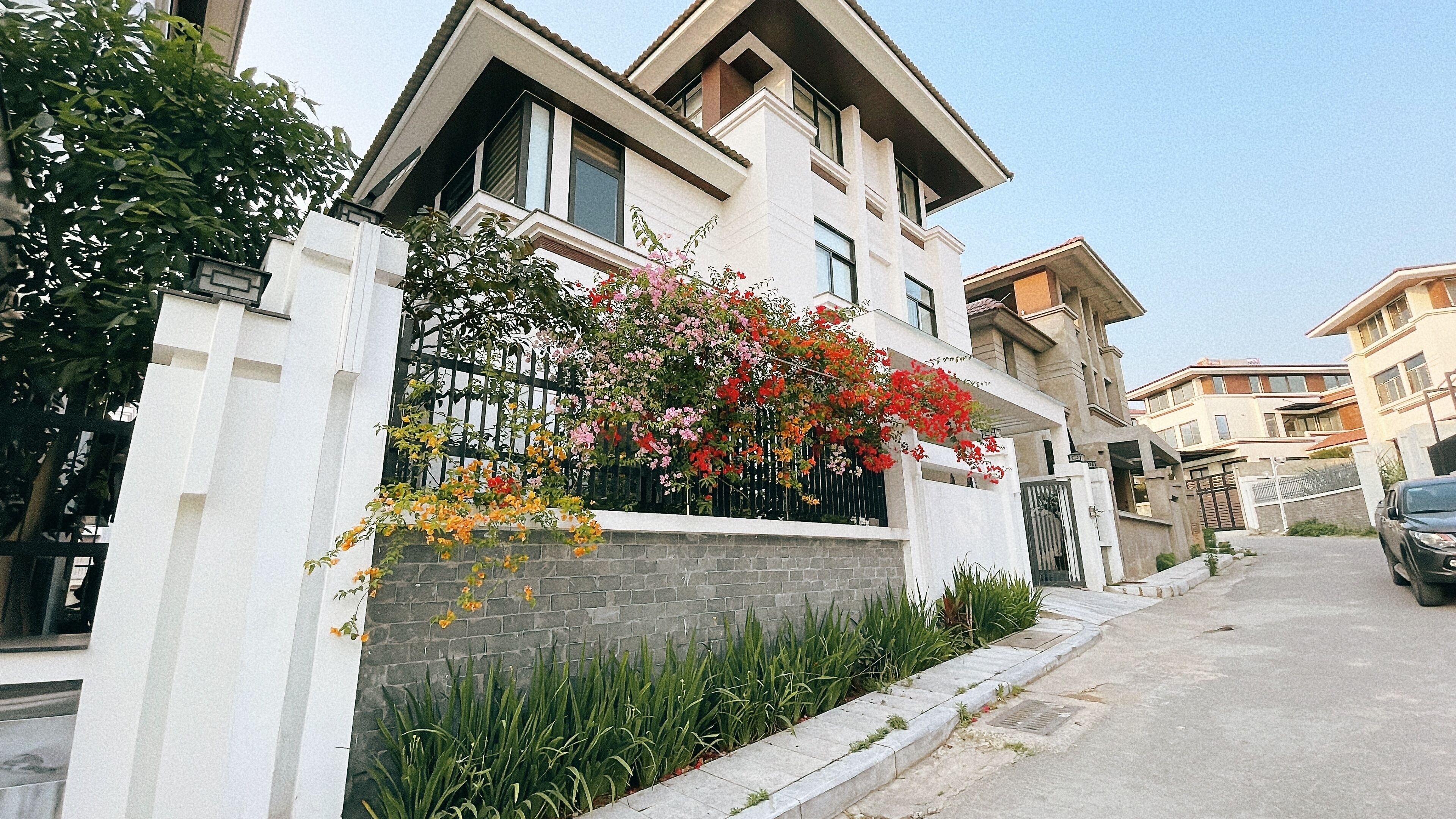A modern two-story house with a stone facade, a balcony with flowering plants, and a gated entrance.