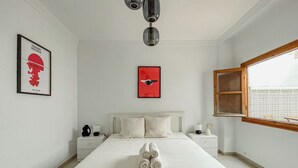 Un dormitorio ordenado con una cama grande, una mesita de noche, una lámpara auxiliar, una obra de arte colgada en la pared y una ventana con marco de madera.