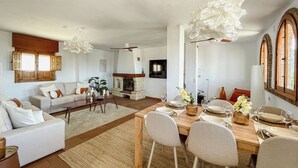 Amplia sala de estar con chimenea, comedor con mesa de madera dispuesta para seis personas y un rincón de descanso con sofá y sillones.