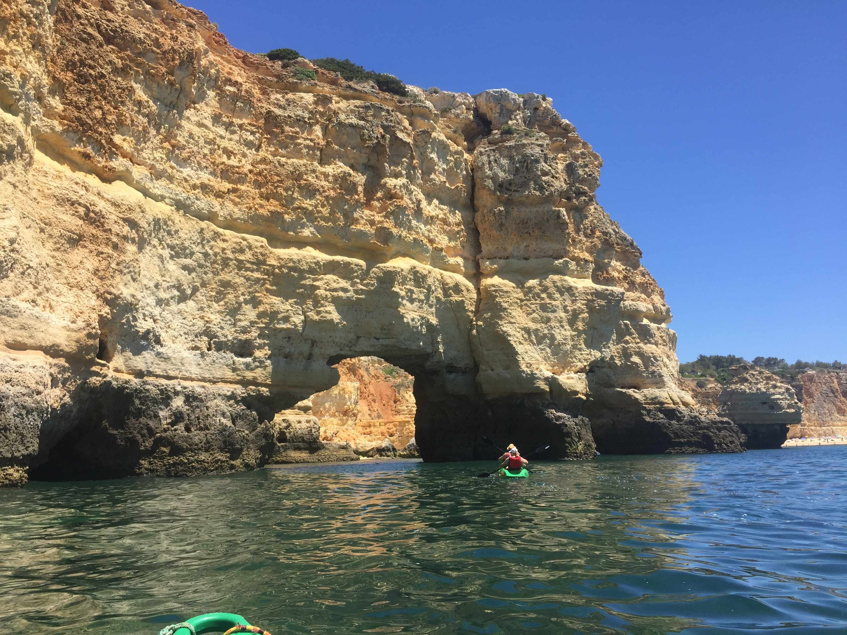 Un kayakiste dans un kayak vert, près d’une grande formation rocheuse surmontée d’une arche naturelle.