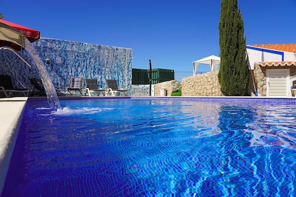 A swimming pool with a water feature, lounge chairs, and a stone wall.