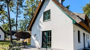 Ein weißes Haus mit einem steilen Satteldach, grünen Fensterläden und einer kleinen Veranda mit Tisch und Stühlen.