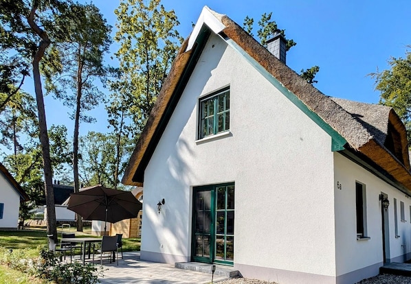 A white house with a steeply pitched roof, green shutters, and a small porch with a table and chairs.
