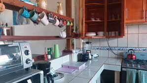 Una cocina con microondas, tostadora, hervidor y licuadora sobre la encimera. Hay varias tazas y una estantería con objetos decorativos.