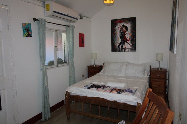 A bedroom with a wooden bed, two bedside tables, a window with curtains, an air conditioner, and a painting on the wall.