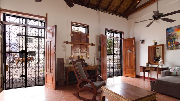 Un salón amplio con una mecedora de madera, una mesa de centro y un sofá. Dos puertas de madera abiertas dan a un balcón con una reja de metal.