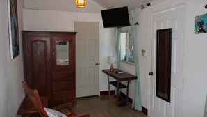 Una habitación con una silla y un armario de madera, una mesa con una lámpara, un televisor fijado en la pared y una puerta.