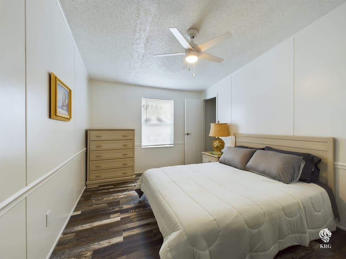 A bedroom with a bed, a ceiling fan, a dresser, a nightstand with a lamp, and a window.