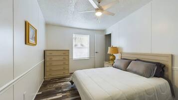 A bedroom with a bed, a ceiling fan, a dresser, a nightstand with a lamp, and a window.
