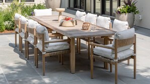 Una mesa de comedor de madera con sillas blancas acolchadas, rodeada de plantas en macetas y un edificio moderno con grandes ventanales al fondo.