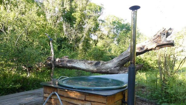A hot tub set up outdoors with a wooden structure, a metal chimney, and a log fire.