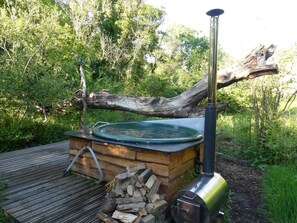 A hot tub set up outdoors with a wooden structure, a metal chimney, and a log fire.