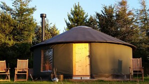 A circular yurt with a door and windows, surrounded by chairs and a kettle on the ground.