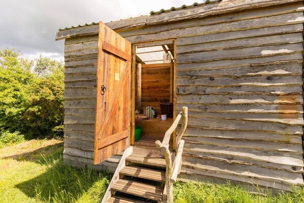 A rustic wooden cabin with a wooden door open, revealing a small interior with a bench and a shelf with books and a starfish.
