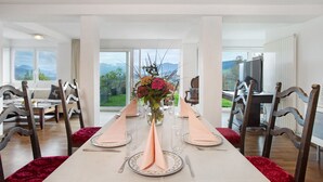 Comedor con una larga mesa dispuesta para cuatro, sillas de madera, un florero con flores y vistas a montañas a través de amplios ventanales.