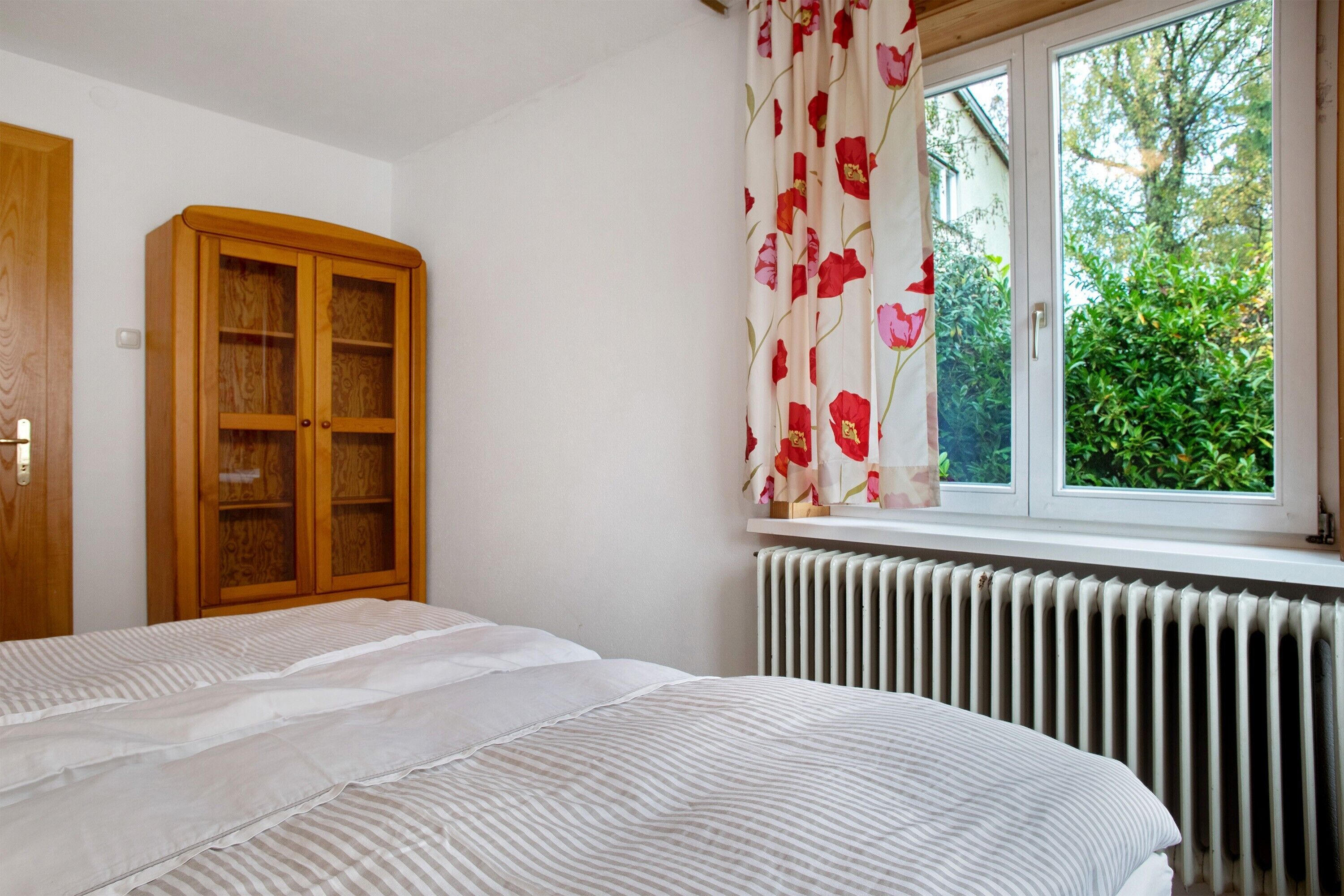 Ein Schlafzimmer mit einem Bett, einem Holzschrank, einem Heizkörper und einem Fenster mit Blick auf Grünflächen.