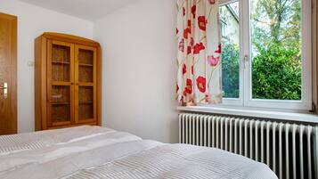 Ein Schlafzimmer mit einem Bett, einem Holzschrank, einem Heizkörper und einem Fenster mit Blick auf Grünflächen.